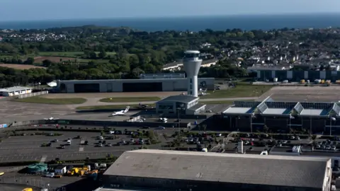 An aerial view of Jersey Airport which shows a large car park to the left with the glass airport building to the right with a triangular glass roof. Behind the airport building is the spherical shaped silver control tower. In the background are houses and trees and behind them is the sea.