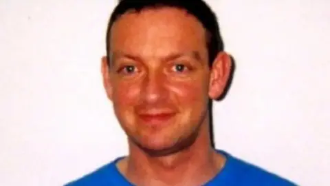 Colin Norris smiling at the camera wearing a blue top, with dark brown hair and stood in front of a white background 