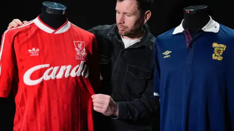 PA Media A Propstore employee looks at Steve Nicol's match-worn red Liverpool v Everton 1989 FA Cup final shirt (est £3,000 - £6,000) alongside his match-worn blue Scotland v England 1988 Rous Cup shirt (est £1,000 - £2,000) during a preview of the upcoming Steve Nicol memorabilia sale