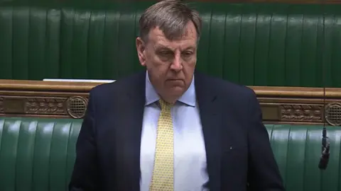 John Whittingdale in the House of Commons, wearing a yellow tie, a blue shirt and a navy blue blazer. The green benches and wood panels are behind him. A black microphone on a cord is hanging from the ceiling.