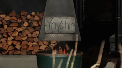 Aberystwyth Town Football Club The sign of Ynshir restaurant's name engraved on a fire pit. There's a fire burning with logs stacked up in the background. 
