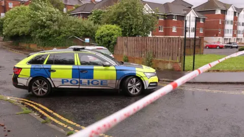 A marked police car is parked behind a red and white police cordon on a small residential street in north Belfast.