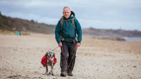 Beat Media/The Kennel Club Nick and Louis walk towards the camera on a wide sandy beach, strewn with pebbles. They are both looking at the camera, and Nick is smiling while holding Louis on the lead.