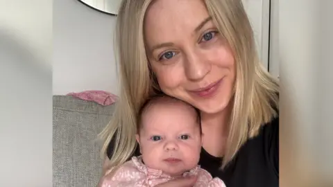 Raegan Thomas A woman with mid-length straight blonde hair wearing a black top sitting on a sofa holding her baby who has a small amount of dark hair and is wearing a pink onesie.