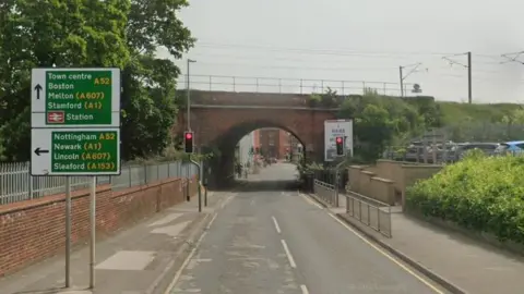 Google Dysart Road, looking towards traffic lights and a red-brick railway bridge. The road surface looks damaged and in the foreground a green sign gives directions to the town centre and other locations, including Boston, Nottingham and the train station.