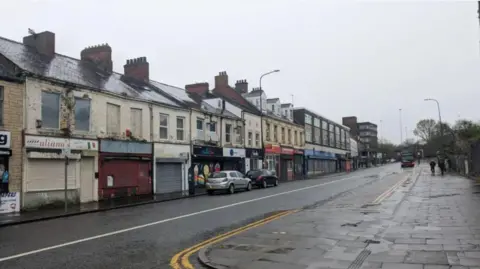 A wet street from the rain on a dark, drizzly say. On the left hand side of the road sits a row of empty shops with shutters boarded up. On the right hand side of the road, two people walk towards a bus stop.