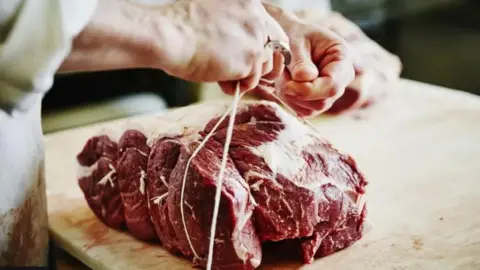 Getty Images A large chunk of red meat sitting on a butchers block. A person wearing a white coat is tying white string around it.