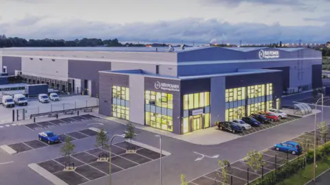 Shaun Flannery The exterior of a huge silver building with a large car park and neatly planted trees