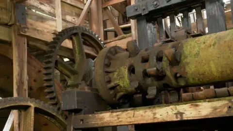 A close up image of part of Tolgus Mill, with big metal wheels and woodwork. Some of the metal looks rusty and is covered in green moss.