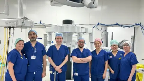 seven people are inside a white operating theatre with white machines in the background. They are all wearing dark blue scrubs and hair nets