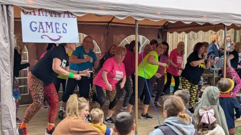 Friends of Eastwood Farm Women dancing on a platform with an audience of families. There is a sign which reads "Carnival games". 