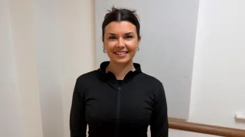 Ballet teacher Grace stands in a dance studio wearing a fitted black zip-up top, with their hands resting by their sides. Behind herare wooden barres mounted on the wall and acoustic panels in a softly lit practice space.