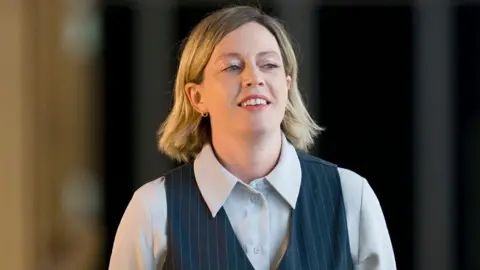 PA Media Jenny Gilruth carries a folder while walking down a corridor in the Scottish parliament. She has shoulder length blonde hair and wears a grey shirt and black pinstripe waistcoat