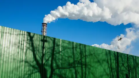 A stock image of chimneys with vapour coming out of the top, behind a green industrial fence.