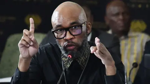 Vusimusi "Cat" Matlala, with the collar of a Fendi shirt sticking out above his jumper, gesticulates as he talks into a microphone.