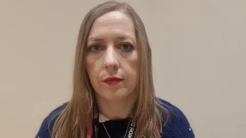 Dr Clodagh Corrigan looks into the camera. She has shoulder length brown hair, blue eyes and is wearing red lipstick. She is wearing a navy jumper.