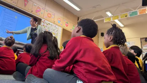 A group of six-year-olds are sitting on the classroom floor, listening to the teacher. She is pointing at a screen displaying the school values. The children are wearing red jumpers and yellow polo-shirts. The walls of the classroom are covered with coloured images and posters to help children with their vocabulary.