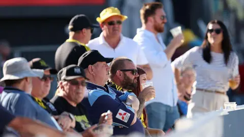 Getty Images A row of cricket fans seated looking to the right of the camera, many are wearing caps and holding beer in their hands. It is a bright, sunny day.