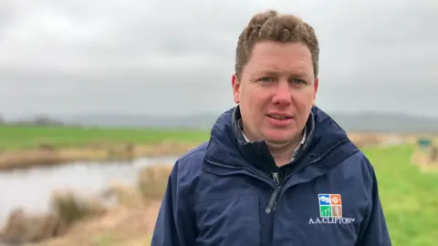 Fiona Irving / BBC A man with curly brown hair looks at the camera. He is wearing a blue jacket with a logo saying "A A Clifton" on it. In the background is a green field and a strip of water.