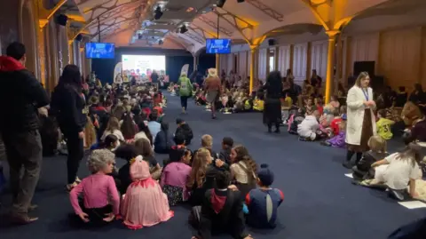 Hundreds of children in fancy dress costumes sit on the floor of a large hall with arched ceilings and warm lighting, facing a stage with screens and book displays at the far end.
