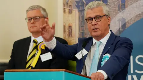Malcolm Offord, who has grey hair and glasses, speaks at a podium with pale blue Reform branding 