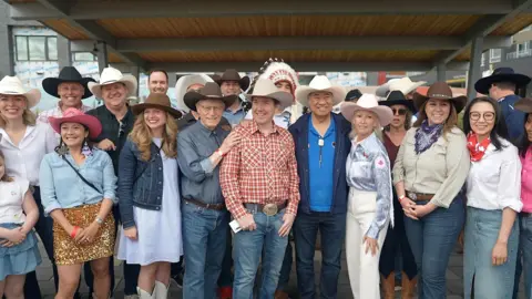 BBC News/Eloise Alanna Corey Hogan, centre, in a red and white plaid shirt, a white cowboy hat, and jeans, smiles and poses with dozens of people, also in western regalia.