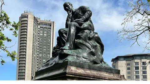 A bronze statue on a marble plinth, surrounded by the tall buildings on Park Lane. The statue depicts Lord Byron seated, with his head resting on his right hand in a pensive mood. 