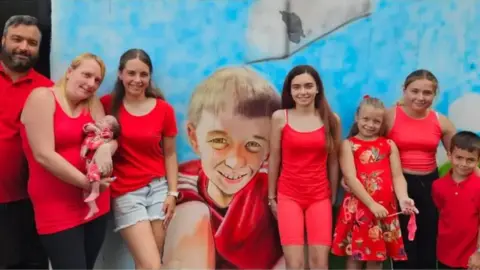 The Dowling family A man with a grey beard and a blonde woman holding a baby, smiling at the camera with their four daughters and son. They're all wearing red and standing in front of a mural depicting the photo of Warren, smiling at the camera and wearing a red shirt