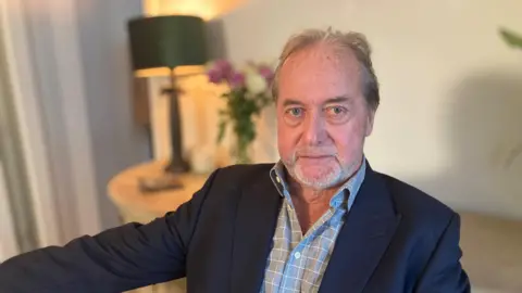 Stephen Bath is sitting in his living room at home, looking straight at the camera with a stern look. He has blue eyes, grey hair and is wearing a grey shirt and navy suit jacket.