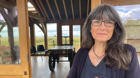 Emma is wearing a black dress and cardigan and glasses and is standing in front of the room where natural burial services are held on her land in Lower tysoe.