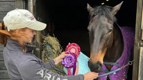 Amy Hilton A woman wearing a cap stood next to a horse holding a purple and pink rosette. The horse is inside a stable door. 