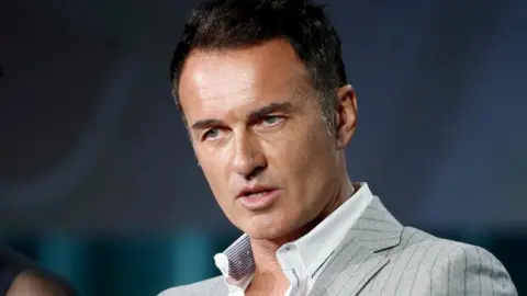 Julian McMahon wearing a white shirt and lined grey blazer in 2016