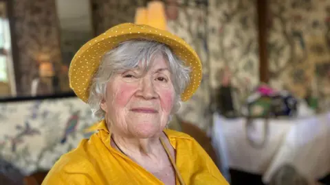 Jeanette, who has grey hair and sparkling brown eyes wears a yellow polka dot hat and yellow linen jacket, as she smiles toward the camera 