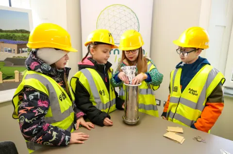 four children in high viz jackets place items in a time capsule