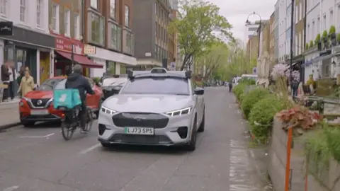A white Ford Mach-E is shown driving down a street in London. A Deliveroo courier is cycling past the left side of car in the opposite direction. The right side of the road is flanked by bushes and flower boxes, which form a barrier between chairs and tables of restaurants' outdoor dining areas on pavements. The BT Tower is visible in the very background.