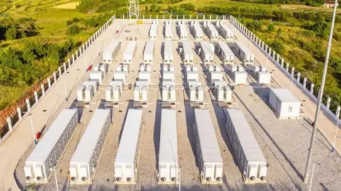 A battery energy storage system, with units spread across a concrete plot in a rural landscape.