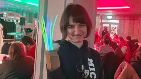 Caring Breaks A young woman with shoulder length brown hair, holding up her arm which has glowsticks attached, at a nightclub with a crowd in the background and neon lights