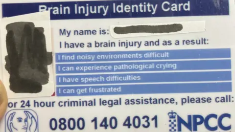 A close-up of a Brain Injury Identity Card. It reads: "My name is: (name is covered over), I find noisy environments difficult, I can experience pathological crying, I have speech difficulties, I can get frustrated." It includes a telephone number for "24-hour criminal legal assistance", and a photograph of the holder is also covered over.