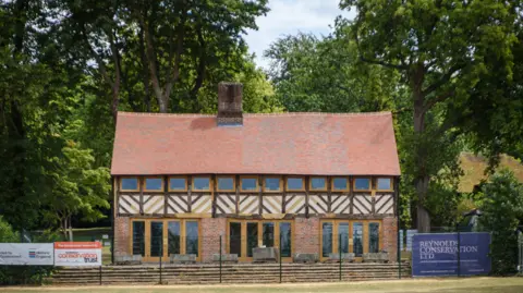 Restored historic timber-framed building with a red tiled roof and brick ground floor, viewed from across a grassy field. Large glass doors line the front façade, with mature trees surrounding the property and conservation project signs displayed along the perimeter fence.