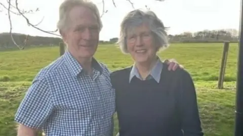 Campbell Welsh dressed in a blue and while gingham short-sleeved shirt with his arm around his wife Sheila, who is wearing a shirt with a collar underneath a navy pullover. They are standing in a field.