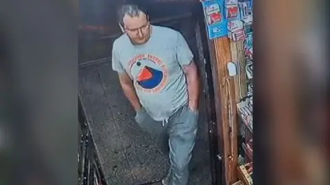 A CCTV image of a Caucasian male entering a shop wearing a grey t-shirt and blue jeans
