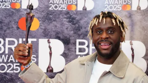 PA Media Myles Smith smiling at the camera with his Rising Star Brit Award. He is wearing a white T-shirt with a beige jacket over the top.