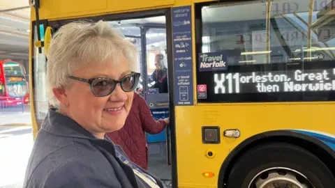 Andrew Turner/BBC Debbie Brown has blonde hair and is wearing tinted glasses. She is smiling, and wearing a blue jacket. Ahead of her in the queue to board the bus is a person in a red quilted jacket. The bus is painted yellow and blue, with the "Travel Norfolk" logo near the door, and the destination board stating "X11; Gorleston, Great Yarmouth then Norwich". A red bus is visible to the left of the image.