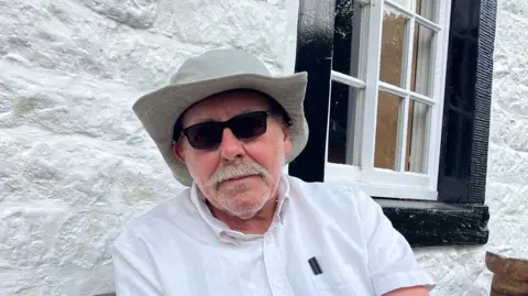 Andy Hopkins is wearing a light cream wide-brimmed sun hat, dark sunglasses, and a white short-sleeved shirt. He sits against a white stone wall beside a black sash window. He has a pocket in his shirt pocket.