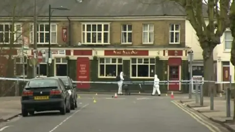 Forensic officers outside The Railway pub. The pub is a two-storey brick building at the end of a terrace of houses. It has a red door and sign that runs across its front.