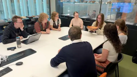 A group of people sit around a table, looking at an object being held. They are a mix of male and female, one holds a bowl in front of the others, another is behind their laptop.

