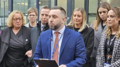 Det Insp Stuart Barson reading a statement from a clipboard. Stephanie's mother and stepfather are stood behind him to his right, and on his left are a number of police liaison officers