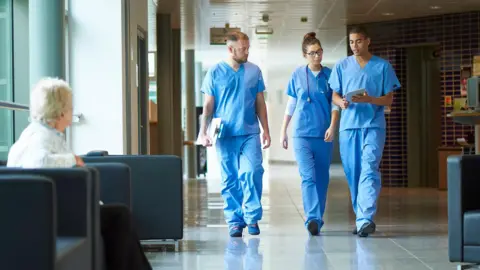 Getty Images Three medics successful bluish scrubs, 2 men and a woman, locomotion on a infirmary corridor. One of nan men is holding a clipboard, nan different immoderate papers and nan female has a stethoscope astir her neck. They are stepping broadside by side, engaged successful conversation. In nan foreground, location is female sitting connected a achromatic cushioned chair. She is turned distant from nan camera, looking towards nan medics.