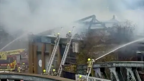 Firefighters on ladders wearing helmets spray water from hoses onto a building. A red fire engine is on the left
