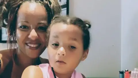 Contributed A selfie style image that is close up of a mum and daughter. The mother is smiling and has light brown hair that has been tied up on top of her head. She is wearing a black halter neck top. Her daughter has light brown hair that has also been tied back. She is wearing a pink top. 
